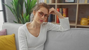 Gözlüklü düşünceli genç bir kadın evdeki bir kanepede oturuyor, zarafeti modern daire dekoruna sıcaklık katıyor..