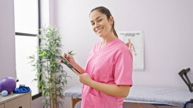 Pembe önlüklü, gülümseyen bir kadın, iyi donanımlı bir rehabilitasyon kliniğinde bir pano tutuyor..