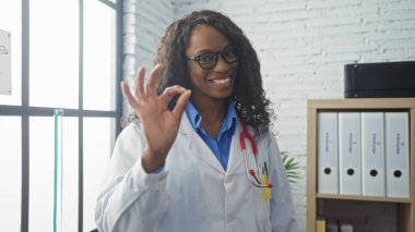 Kıvırcık saçlı genç Afro-Amerikalı kadın doktor hastane kliniğinde gözlük ve steteskop takarak iyi bir jest yapıyor..