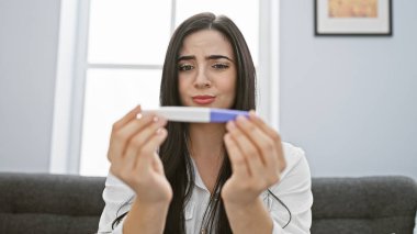 Modern bir oturma odasında hamilelik testi isteyen endişeli, genç bir İspanyol kadın..