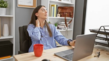 Modern ofis ortamında boynu ağrıyan genç İspanyol kadın, dizüstü bilgisayarda yazıyor.