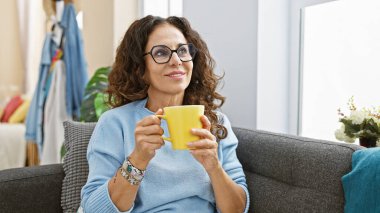 Sarı bir kupa tutarken gülümseyen kıvırcık saçlı ve gözlüklü olgun bir kadın, gri bir koltukta oturuyor.