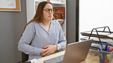 Ofis masasında rahatsız bir kadın, dizüstü bilgisayarla çalışırken karnını tutuyor..