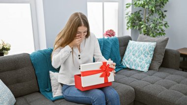 Şaşırmış genç bir kadın, elinde duygu dolu kırmızı bir hediye kutusu tutuyordu, gri bir kanepede oturuyordu..