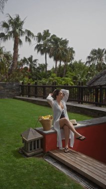 Genç bir kadın lüks bir bali tatil köyünde dinleniyor, tropikal yeşillikler ve şık açık hava dekorları arasında huzur yayıyor..