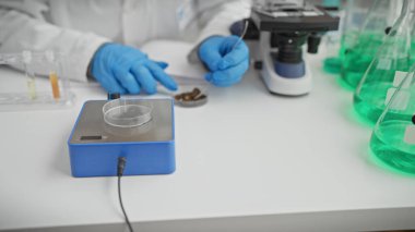 Eldiven giyen bir bilimadamı, mikroskop ve yeşil sıvı şişeleriyle donatılmış bir laboratuvarda örnekleri inceliyor..