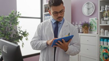 Laboratuvar önlüğü giyen, sakallı ve gözlüklü genç bir İspanyol adam, iyi donanımlı bir eczanede bir tablet kullanıyor..