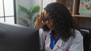 Kıvırcık saçlı ve gözlüklü genç bir Afro-Amerikan kadın doktor klinik masasında baş ağrısı çekiyor..