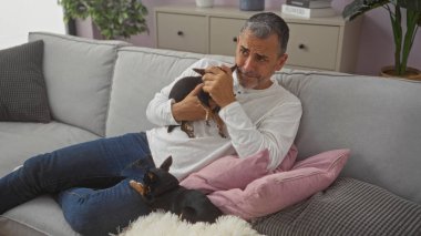 Orta yaşlı İspanyol bir adam, oturma odasında iki küçük chihuahuayla dinleniyor, bir tanesini kucaklıyor, kapalı bir koltukta rahat bir şekilde oturuyor..