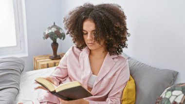 Genç, kıvırcık saçlı, rahat bir yatak odasında kitap okuyan, rahat ve rahat görünen İspanyol bir kadın..