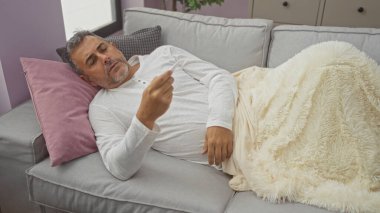 Evdeki kanepede dinlenen gri saçlı orta yaşlı bir adam modern bir oturma odasında rahat bir battaniyeye sarılı bir termometreye bakıyor..