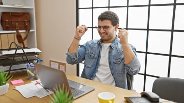 A jubilant young man with glasses celebrates in a modern office, exhibiting a casual style with his denim jacket and joyful expression. clipart