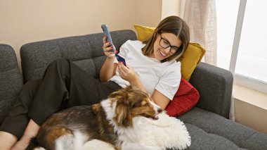 Genç bir İspanyol kadın güzel köpeğiyle evde rahatlıyor, cep telefonu tutarken gülümsüyor.