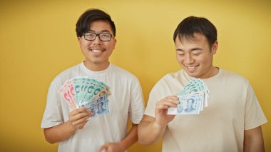 İki Asyalı gülümsüyor ve sarı bir duvara Çin Yuanı notaları tutuyorlar.