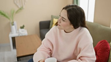 Rahat bir oturma odasında kupa tutan düşünceli genç bir kadın.