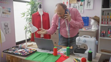 Olgun bir terzi telefon görüşmesi yapıyor ve atölyede çalışıyor. Etrafı dikiş aksesuarları, kumaş ve mankenlerle çevrili..