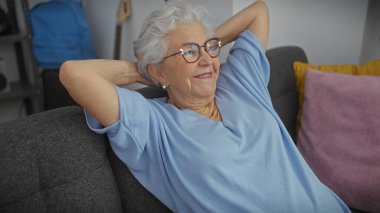 Rahat, gülümseyen, gözlüklü yaşlı bir kadın rahat bir oturma odasında gri bir koltukta oturuyor..
