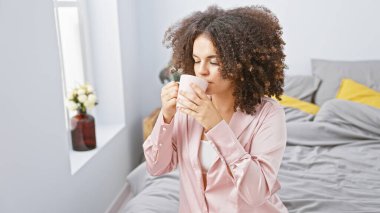 Kıvırcık saçlı genç İspanyol kadın rahat bir yatak odasında kahvenin tadını çıkarıyor.