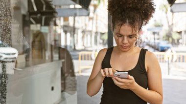 Kıvırcık saçlı genç bir İspanyol kadın güneşli bir şehir sokağında akıllı telefon kullanıyor..