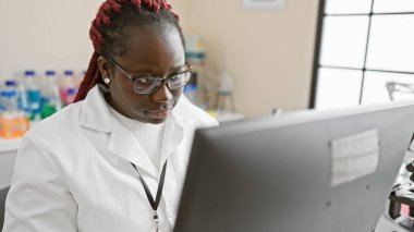 Örgü ören Afrikalı Amerikalı kadın bilim adamı laboratuvar ortamındaki bir bilgisayarda dikkatle çalışıyor..