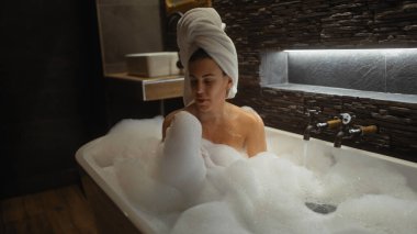 Genç bir kadın, kafasında havluyla modern bir banyoda rahatlatıcı bir köpük banyosunun keyfini çıkarıyor..