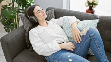 Dinlenmiş İspanyol kadın modern bir oturma odasında, kulaklıkla müzik dinliyor..