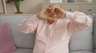 Kafkasyalı yaşlı bir adam, sıcak bir oturma odasında kanepede otururken elleriyle kalp şekli yapıyor..