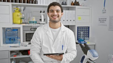 Sakallı, laboratuvar önlüğü giyen ve elinde tablet olan gülümseyen yetişkin bir adam, iyi donanımlı bir laboratuarda duruyor..