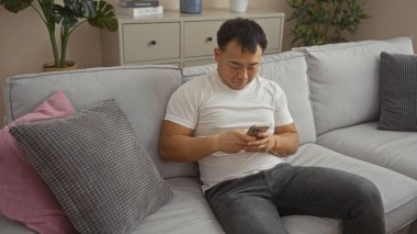 Oturma odasında oturan genç Çinli bir adam, arka planda modern ve rahat bir dairesi olan bir koltukta otururken telefon kullanıyor..