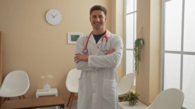 Boynunda steteskop olan beyaz önlüklü genç İspanyol erkek doktor, kolları çapraz bir klinik bekleme odasında güvenle duruyor, profesyonelliği ve arkadaşlığı iletiyor..