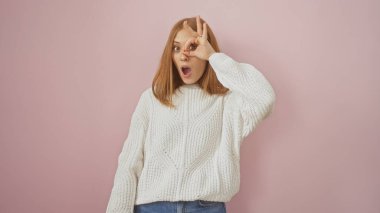 Beyaz kazak giyen neşeli, genç bir kızıl kadın pembe arka plana karşı eğlenceli bir jest yapıyor..