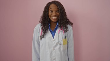 Kıvırcık saçlı genç Afrikalı Amerikalı kadın doktor izole edilmiş pembe arka planda kameraya gülümsüyor..