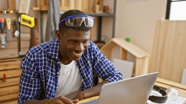 Gülümseyen Afrikalı bir adam ahşap atölyesinde dizüstü bilgisayar kullanıyor..
