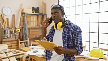 Marangozluk atölyesinde pano tutan Afrikalı bir adam telefonda konuşuyor.