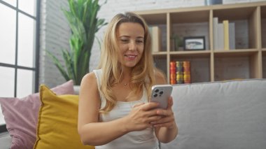 Sarışın bir kadın, rahat bir oturma odasında akıllı telefon kullanıyor. Rahatlık ve modernlik yayıyor..