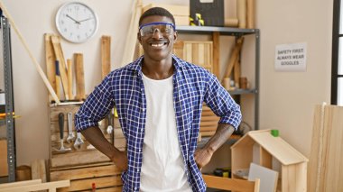 Koruyucu gözlüklü gülümseyen Afrikalı adam bir marangozhane atölyesinde güvenle duruyor.