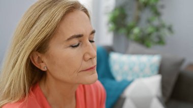 Oturma odasında tefekkür eden beyaz bir kadın, olgunluğu ve huzuru somutlaştırıyor..