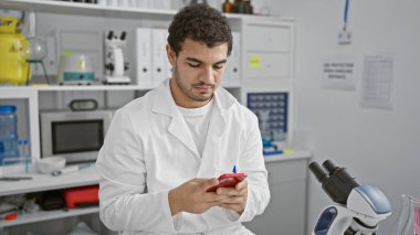 Laboratuvar ortamında akıllı telefon kullanan, beyaz önlük giyen ve cihaza odaklanmış sakallı bir adam.