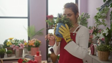 Çiçekçide genç bir kadın çeşitli renkli çiçeklerle çevrili bir bitkiyi nazikçe kokluyor..