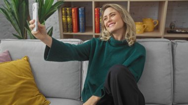 Gülümseyen sarışın kadın rahat bir oturma odasında selfie çekiyor.