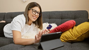 Gözlüklü İspanyol kadın oturma odasında online alışveriş için tablet ve kredi kartı kullanıyor.