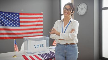 Orta yaşlı, gülümseyen İspanyol bir kadın, kollarını çapraz yapıp Amerikan demokrasisini sembolize eden bir Amerikan bayrağı ve oy sandığının yanında duruyor..