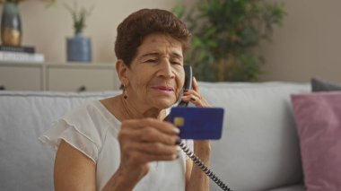 Kısa saçlı, yaşlı bir İspanyol kadın oturma odasında telefonla konuşurken elinde mavi bir kredi kartı tutuyor ve evde tipik bir ev ortamını canlandırıyor..