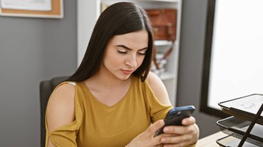 Çekici genç İspanyol kadın modern ofis ortamında akıllı telefon kullanıyor..