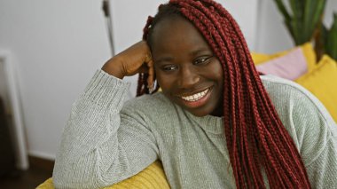 Kırmızı örgülü neşeli bir Afrikalı kadın evinde oturuyor, rahat bir iç mekânda sıcacık bir güzellik ve sıcaklık yayıyor..