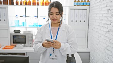 Beyaz önlüklü genç bir İspanyol kadın laboratuvar teknisyeni, etrafı ekipmanlarla çevrili, parlak bir laboratuvar ortamında akıllı bir telefonla mesajlar yolluyor..