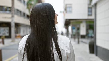 Uzun esmer saçlı, beyaz ceketli genç bir İspanyol kadın şehir sokağına bakıyor..