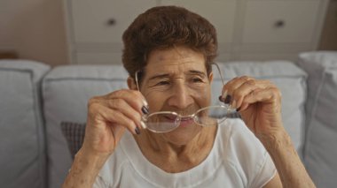Kısa saçlı, yaşlı bir İspanyol kadın. Gözlüklerini kapalı bir oturma odasında ayarlıyor..