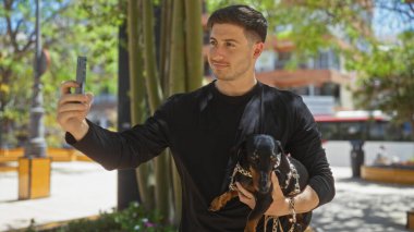 Genç İspanyol bir adam güneşli bir günde tekel köpeğini tutarken şehir parkında selfie çekiyor..