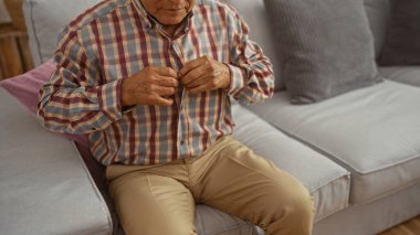 Yaşlı bir adam, rahat bir oturma odasında gri bir kanepede otururken ekose gömleğini ilikliyor..